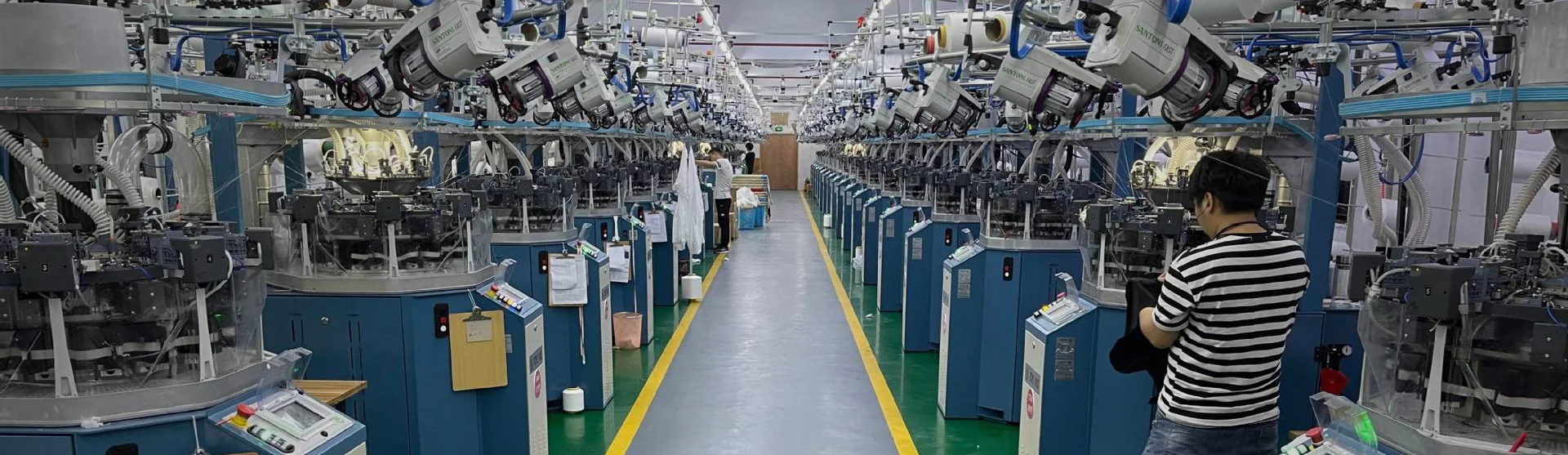 The knitting hall seen down the main aisle, circular knitting machines on both sides and an operator at the near machine