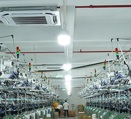 A long hall of circular knitting machines with overhead yarn creels and workers further down the aisle